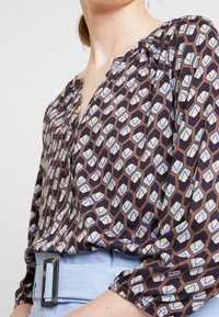 Blusa marrone e blu con motivo a V, spalle arricciate e maniche lunghe, abbinata a una cintura azzurra chiara con fibbia rettangolare.