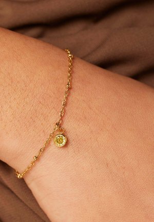Gold bracelet featuring a small round yellow gem pendant, with a delicate chain and a smooth, shiny surface. Textured skin is visible beneath.