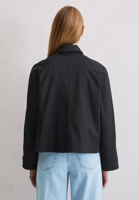 Czarna kurtka o luźnym kroju, ze sztywnym kołnierzem i długimi rękawami, zestawiona z jasnoniebieskimi dżinsami, widziana od tyłu. Gładka faktura.