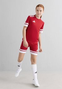 Rød sportsdrakt med hvite striper, kombinert med hvite knesokker og fotballsko. Adidas-merke på drakten og skoene.