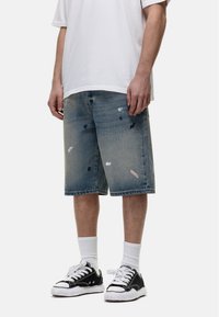 Denimshorts in vervaagd blauw met verfsplatterdetails, losse fit en knielange ontwerpen, gecombineerd met zwarte sneakers en witte sokken.