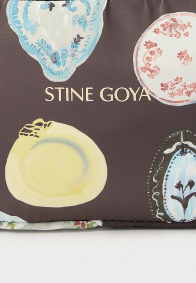 Σκούρο ύφασμα με ζωγραφισμένα σχέδια κεραμικών πιάτων σε μπλε, κίτρινο, κόκκινο και μαύρο, που περιβάλλουν το κείμενο "STINE GOYA" σε χρώμα κρεμ.