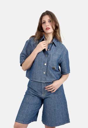 Donna con capelli castano chiaro che indossa una camicia di jeans a maniche corte abbinata e pantaloncini di jeans al ginocchio su sfondo bianco semplice.