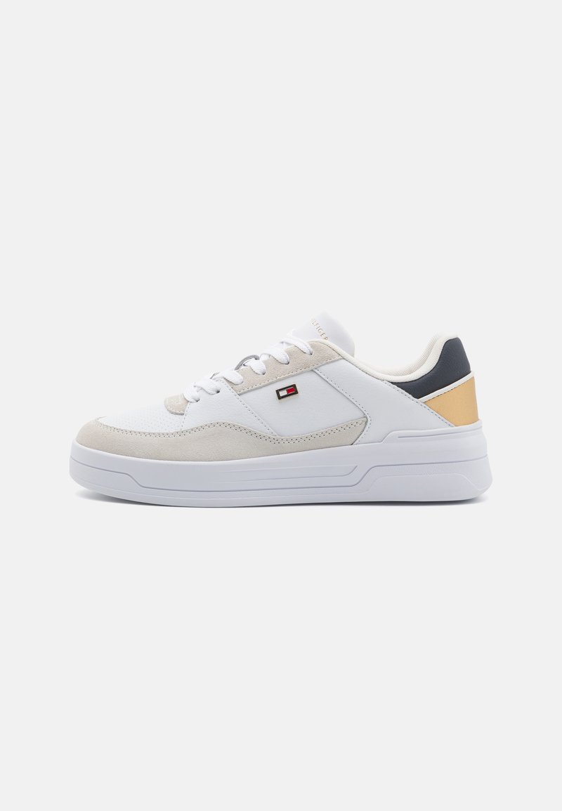 Fehér sneaker szürke velúr részletekkel és bézs hátpanellel. Laposszíjú, párnázott talppal rendelkezik, és hímzett logó található az oldalon.