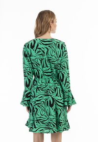 Grünes Kleid mit schwarzem Tigerstreifenmuster, langen Ärmeln, ausgestellten Manschetten und elastischem Bund. Der Stoff wirkt glatt mit leichtem Glanz.