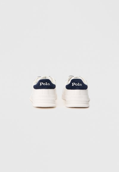 Ténis brancos com acentos em azul-marinho, apresentando um topo liso, sola em borracha e a marca "Polo" no calcanhar e na língua.