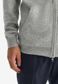 Grå zip-up cardigan i en mjuk, fluffig textur, med en framficka och ribbade ärmslut, i kombination med mörka byxor.