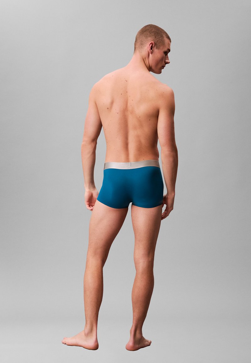 Boxers pour hommes couleur sarcelle avec une ceinture argentée, présentant une texture lisse et une coupe ajustée, vus de dos.