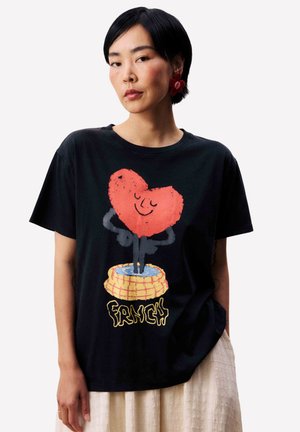 Femme portant un T-shirt noir avec un cœur rouge souriant sortant d'un panier jaune marqué "FRANÇAIS", assorti à une jupe beige et des boucles d'oreilles en forme de fleurs rouges.