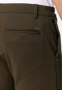 Pantaloni verde oliva con una texture liscia, dotati di una tasca posteriore con un bottone marrone e un piccolo particolare in metallo.