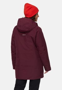 Osoba w bordowej kurtce z kapturem z logo "MAMMUT" na plecach i jasnoczerwonym czapce, zwrócona bokiem na jednolitym tle.