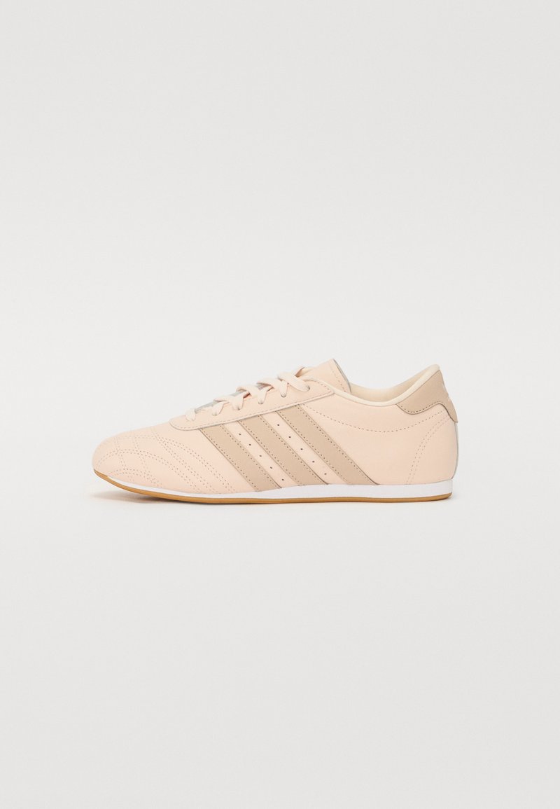 Zapatilla deportiva de color rosa claro con un upper de cuero liso, tres rayas beige, punta redondeada, suela blanca plana y un mínimo de perforaciones decorativas.