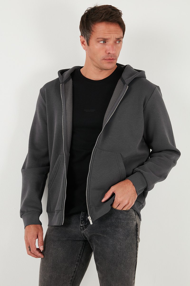 Grå zip-up hoodie gjord av mjukt material, med huva, framfickor och en slät dragkedja. Bärs över en svart t-shirt och mörka jeans.