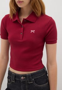 Rotes, geripptes Poloshirt mit Kragen, ausgestattet mit drei Knöpfen und einer kleinen rosa Schleife, die auf der linken Brust gestickt ist.