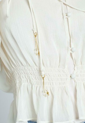 Blusa bianca con un tessuto testurizzato, caratterizzata da una vita arricciata, bottoni rotondi e dettagli dorati con perline pendenti ai lati.
