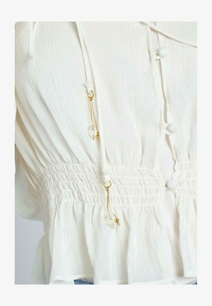 Blusa bianca con un tessuto testurizzato, caratterizzata da una vita arricciata, bottoni rotondi e dettagli dorati con perline pendenti ai lati.