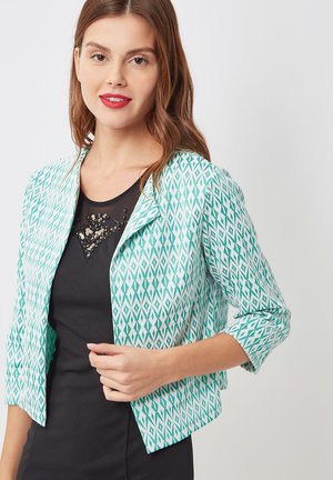 Mujer joven con una blazer de patrón de rombos en turquesa y blanco sobre un vestido negro con detalle de lentejuelas en el pecho, sonriendo suavemente.