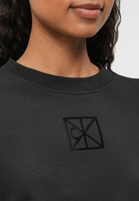 Μαύρο φούτερ με στρογγυλή λαιμόκοψη, υφασμάτινη υφή και έντονο μαύρο λογότυπο στην αριστερή πλευρά του στήθους.