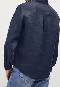 Marinblå linneskjorta med knäppte ärmslut, en ryggholk och en rundad fåll. Tyget har en subtil struktur och ett lätt utseende.