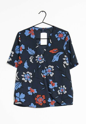 Blouse bleu marine à manches courtes avec un motif floral rouge, blanc et bleu, suspendue sur un cintre noir sur fond blanc.