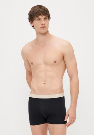 Cajones negros con una cinturilla beige, diseño ajustado, hechos de una tela suave. El modelo está sin camiseta, de pie en un entorno neutral.