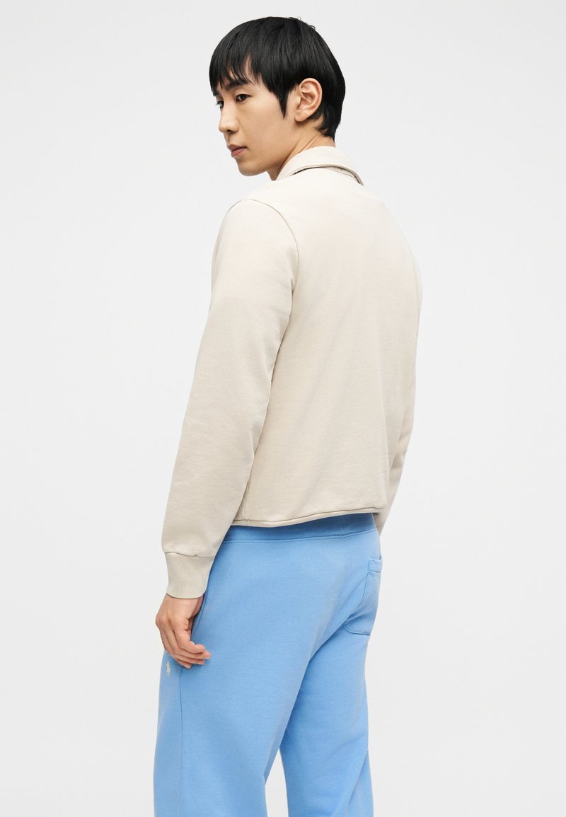 Jeune homme de profil portant une veste beige et un pantalon de survêtement bleu clair sur un fond blanc uni.