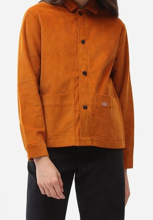 Kvinde iført en brændt orange fløjlsjakke med sorte knapper og forlommer, kombineret med sorte fløjlsbukser.