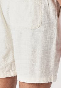 Pantalones cortos de color claro con textura y un patrón de cuadrícula, que cuentan con un bolsillo trasero y un dobladillo recto. El material parece ser tejido y transpirable.