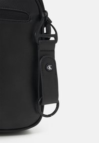Angolo di una borsa di pelle nera con tracolla attaccata, caratterizzata da un anello metallico e dettagli con il logo "cK" di Calvin Klein.