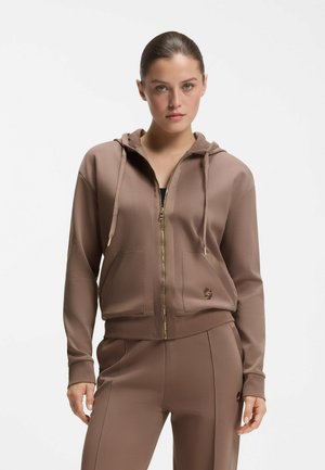 Kobieta ubrana w brązową bluzę z zamkiem na zamek błyskawiczny z sznurkami i dopasowane spodnie w tym samym kolorze, stojąca na tle białego, jednolitego tła.