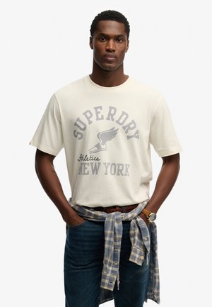 Cremiges Kurzarm-T-Shirt mit grauem Grafikdruck, einschließlich "SUPERDRY" und Flügel-Logo, getragen mit blauen Jeans und karierter Hemd, das in der Taille gebunden ist.
