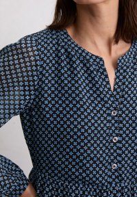 Navy blouse met een bloemendessin in lichtblauw. Heeft een ronde halslijn, een knoopsluiting aan de voorkant en een gather-zoom met lange mouwen.