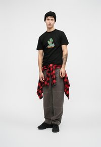 Melna t-kreklu ar zaļu grafiku, pelēkus brīvus bikses, sarkanu un melnu kvadrātains flaneli, kas sasietas ap vidukli, un melnas kedas.