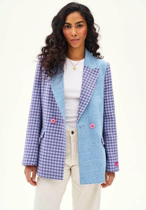 Kobieta w marynarce w fioletowo-niebieską pepitkę z różowymi guzikami, białej koszuli, beżowych spodniach i złotym wisiorze na białym tle.