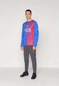 Pitkähihaisen urheilupaidan väri liukuu sinisestä punaiseen, jossa on "QATAR AIRWAYS" -logo, yhdistettynä harmaisiin urheiluhousuihin ja mustiin kenkiin.