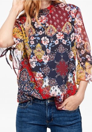 Nainen, joka käyttää moniväristä, kukka- ja paisley-kuvioista puseroa, jossa on läpikuultavat 3/4-hihapunosolmut, yhdistettynä sinisiin farkkuihin.