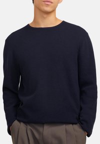 Mann in einem strukturierten, navyblauen Langarmpullover mit braunen, gefalteten Hosen, die Hände in den Taschen, gezeigt vom Brustbereich bis zur Hüfte.