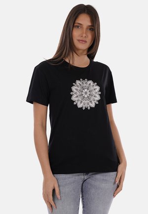 Donna che indossa una t-shirt nera con un complesso disegno mandala bianco al centro, abbinata a jeans azzurri, in piedi davanti a uno sfondo neutro.