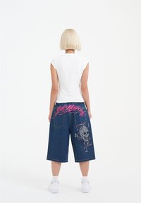 Denim culottes featuring a skull and snake embroidery in gray and pink. Reversible design with a white fitted top, shown from behind.