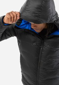 Personne ajustant la capuche d'une veste en duvet noire avec doublure bleue, couvrant partiellement son visage, contre un fond clair uni.