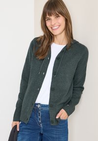Dunkelgrüner Pullover mit Kapuze, vorderen Knöpfen, weichem Material, langen Ärmeln und lässigem Design, kombiniert mit einem weißen Hemd und blauen Jeans.