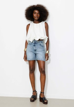 Femme avec une coiffure afro naturelle portant un chemisier blanc sans manches, un short en jean clair, des bracelets dorés et des sandales à plateforme noires, debout contre un mur blanc.