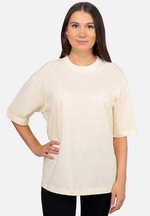 Vrouw met lang bruin haar die een loszittend crèmekleurig T-shirt met korte mouwen draagt, staand met één hand op de heup tegen een effen achtergrond.