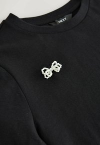 Czarny bawełniany t-shirt z białą, koralikową ozdobą w kształcie kokardy na piersi, podkreślający teksturalny design z gładkim wykończeniem materiału.