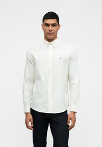 Camisa branca de manga longa com botão, com um pequeno logótipo no lado esquerdo do peito, combinada com jeans escuros, num fundo branco liso.