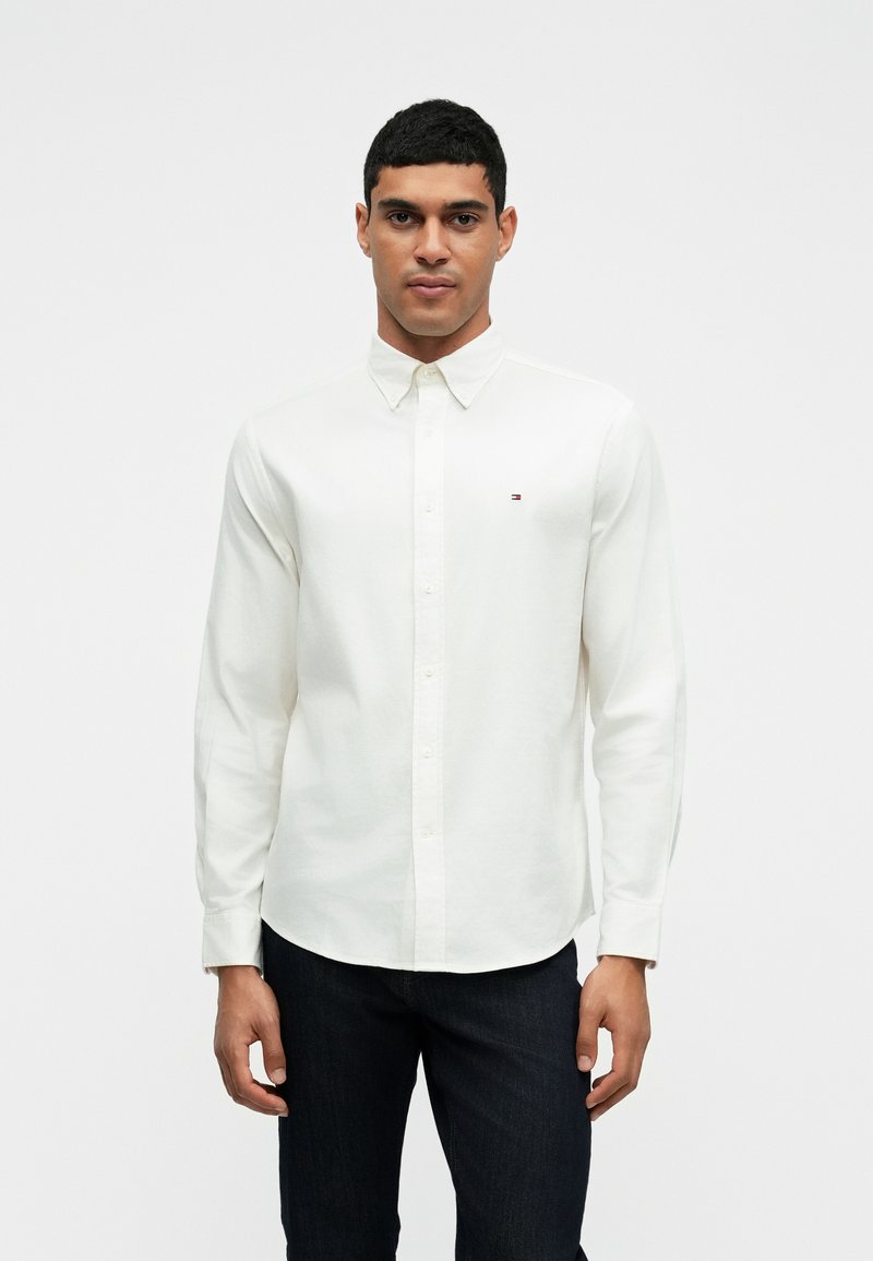 Camisa branca de manga longa com botão, com um pequeno logótipo no lado esquerdo do peito, combinada com jeans escuros, num fundo branco liso.