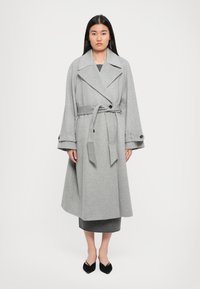 Grå ulltrenchcoat med breda ärmar, stor krage och matchande bälte. Har knapppdetaljer på ärmsluten.