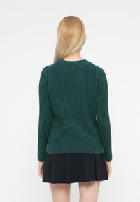 Pull en maille côtelée verte à manches longues, avec un col rond et une taille ajustée, associé à une jupe plissée noire.