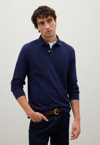 Junger Mann mit lockigem Haar, der ein navyblaues langärmliges Poloshirt, ein weißes Unterhemd, dunkle Jeans und einen braunen geflochtenen Gürtel trägt, steht mit einer Hand in der Tasche.