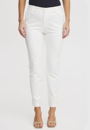 Femme portant un pantalon blanc ajusté et des talons ouverts beiges, debout devant un fond uni.